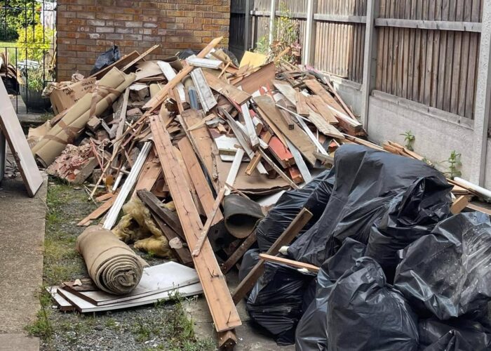 A pile of garden rubbish, including bin bags, leaves, branches, grass cuttings, and old wooden planks, stacked near a fence, ready for collection.