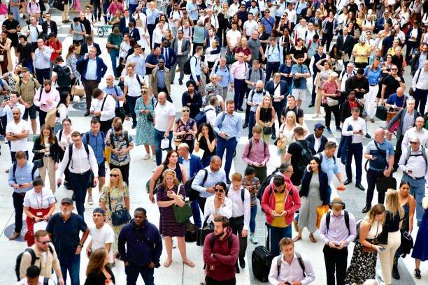 A large crowd of people gathered outdoors, standing close together. Some individuals are talking, while others are walking or looking around.